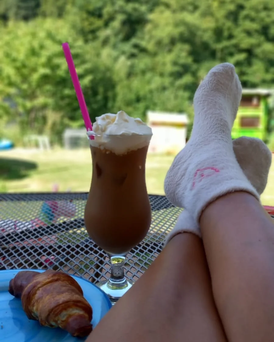 Ein Glas Eiskaffee mit Sahne und pinkem Strohhalm neben einem Croissant auf einem blauen Teller und hochgelegten Füßen in weißen Socken im sonnigen Garten.